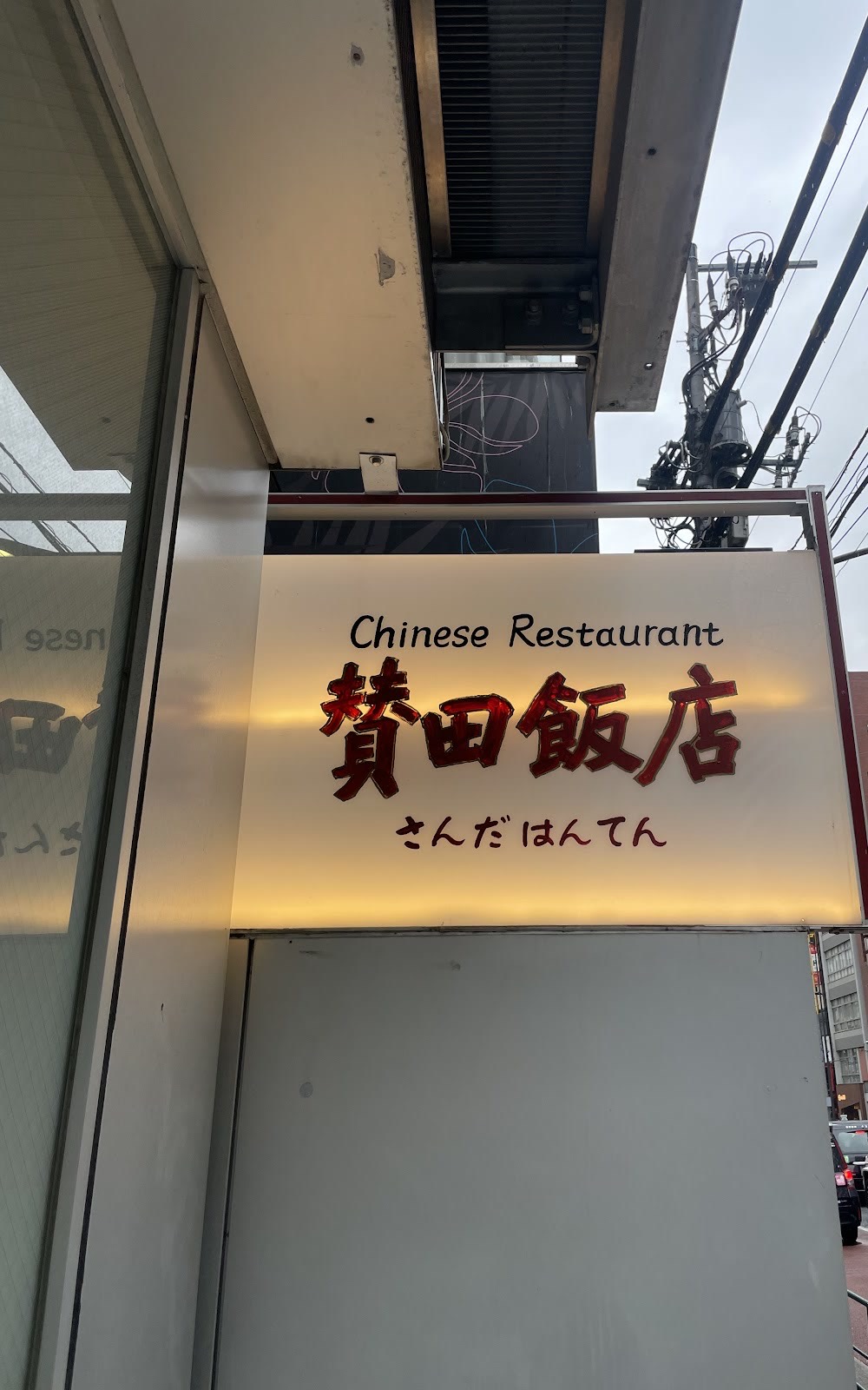 白い行灯看板。「Chinese Restaurant」の黒い文字の下に赤い太字の「賛田飯店」、その下に小さく「さんだはんてん」。上に庇と電線、曇り空。