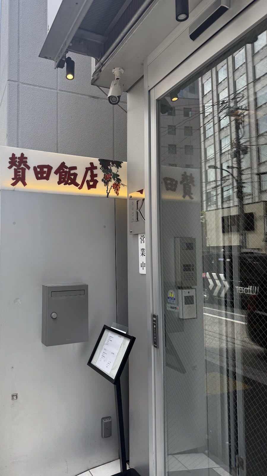 白い外壁に赤い文字の「賛田飯店」の看板と葡萄の絵。ガラスの引き戸、インターホン、黒いメニュースタンド、小さな「営業中」の札。