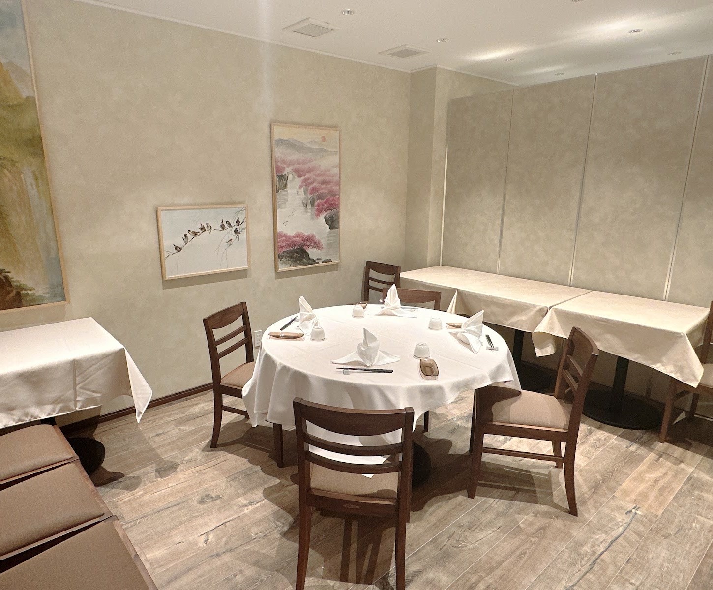 山水画や枝に止まる鳥、桃色の花の絵が掛かる部屋。白いクロスの円卓と壁ぎわのテーブル、淡い色の間仕切り。