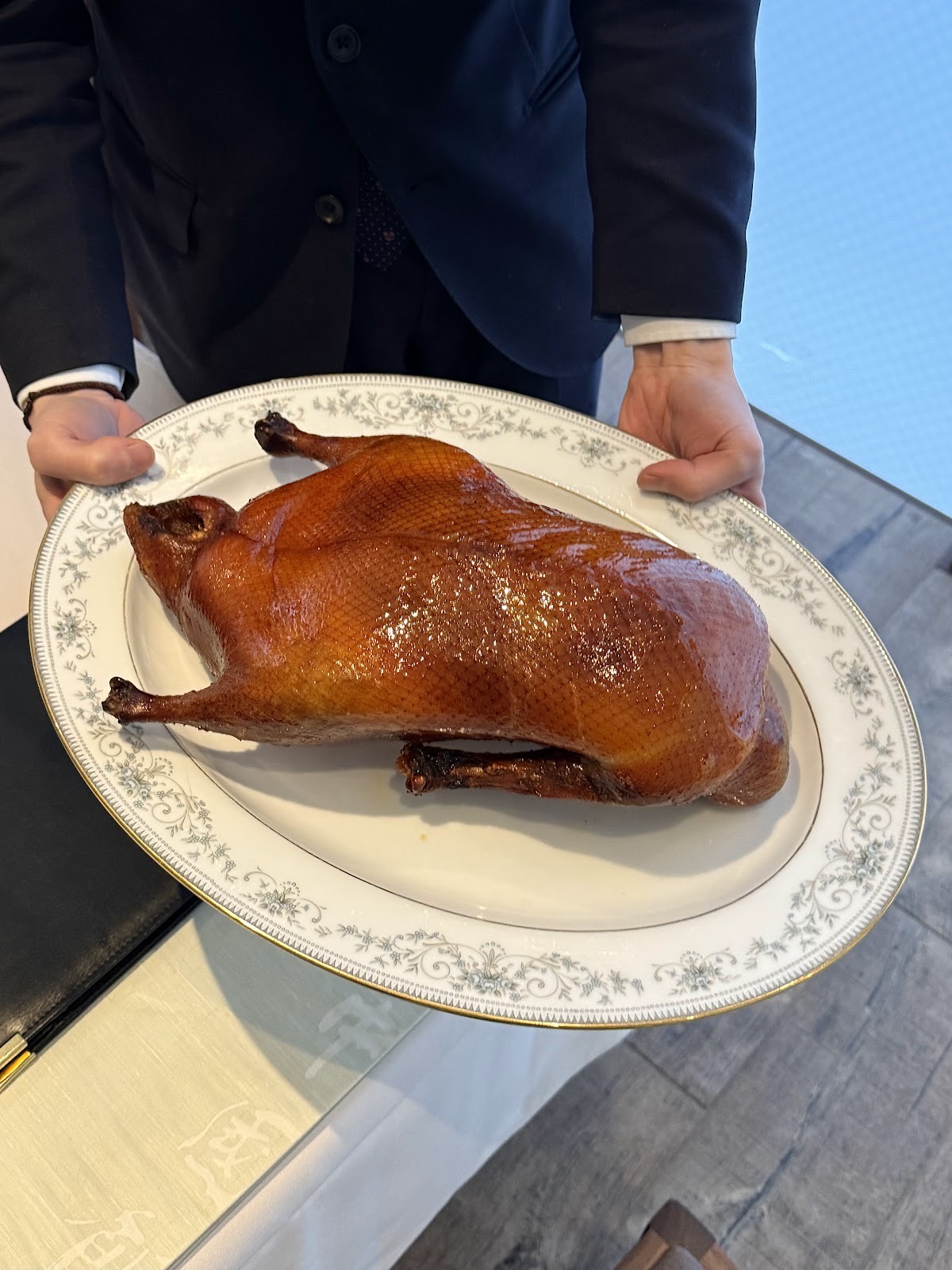 紺のスーツのスタッフが両手で持つ、花柄の縁の楕円の皿に載った艶のある飴色の丸ごと一羽のローストダック。