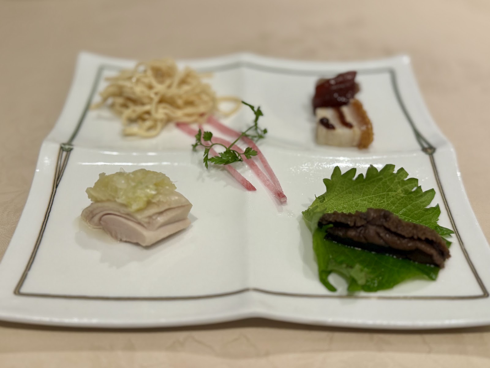 四つに仕切られた白い皿の前菜四種。もやしを添えた細切り、照りのある一切れ、淡い色の薄切り肉、大葉に載せた黒い一品。