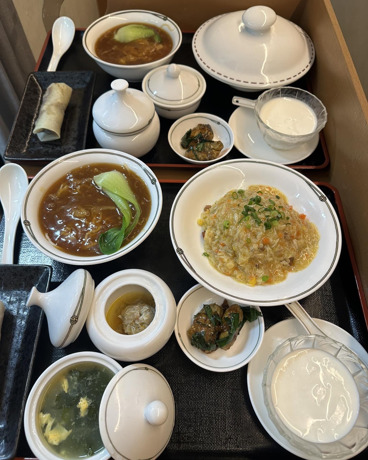 黒いトレーに並ぶランチセットを真上から。青菜の入ったとろみのある茶色いスープ、淡いあんのかかった炒飯、蓋付きの器、黒い皿の薄皮の包み、青菜の小皿、白いデザート、卵スープ。
